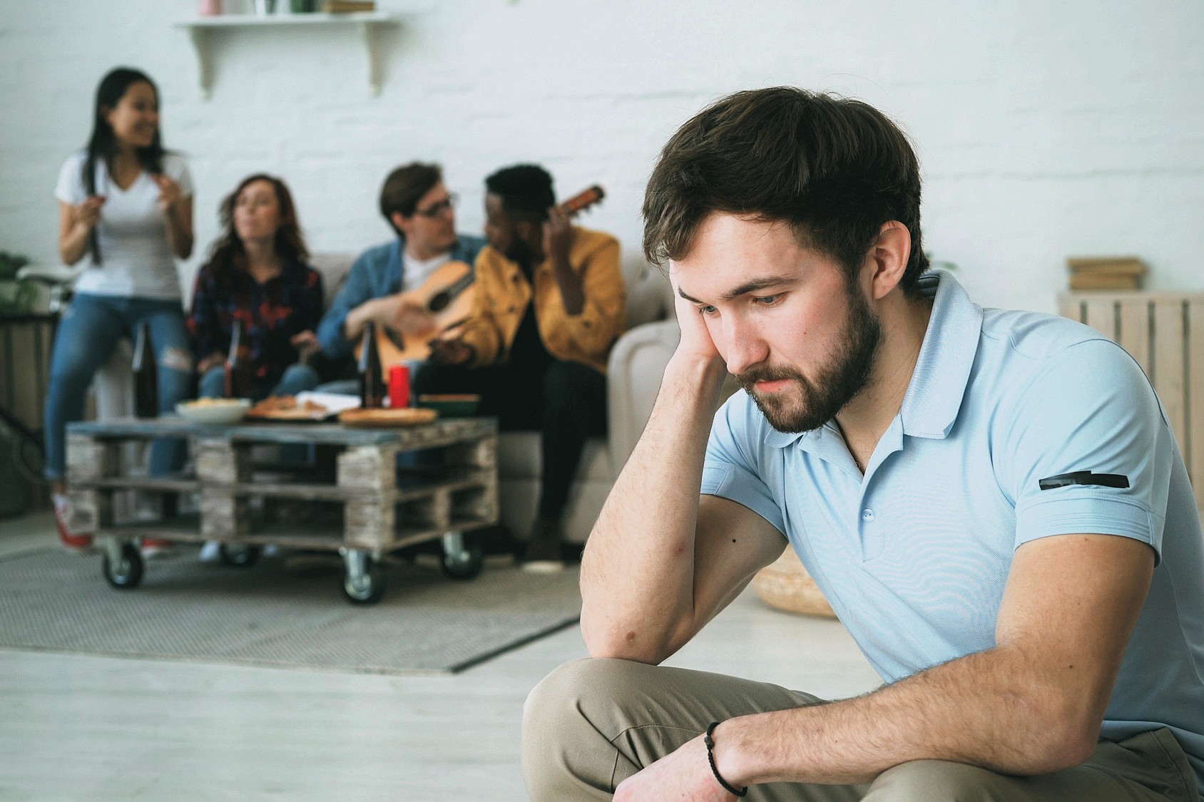 A man sits apart, looking sad and thoughtful, while a group of four people in the background sing, play guitar, and enjoy each other's company in a cozy living room.