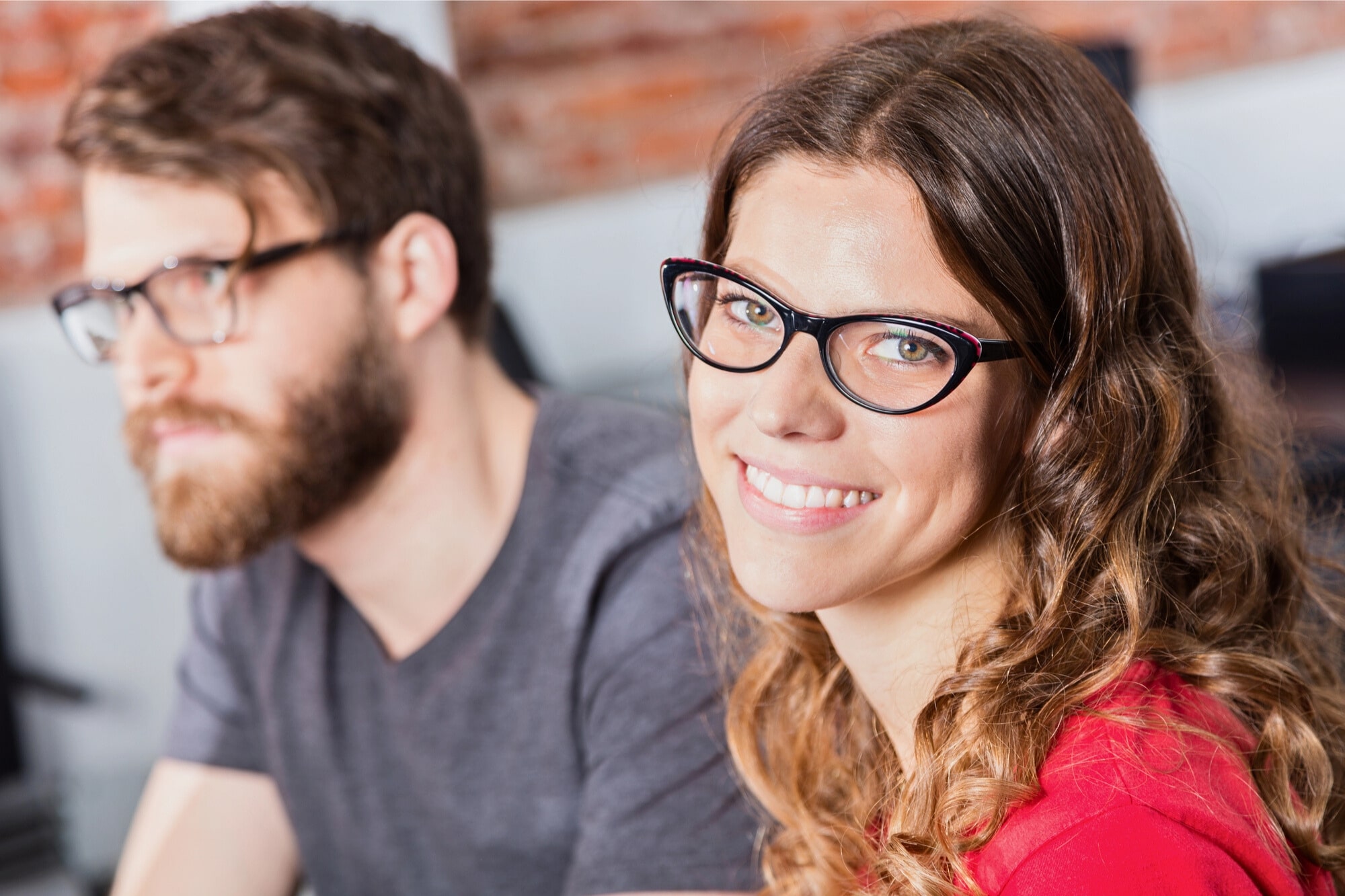A woman with long wavy hair and glasses smiles at the camera, while a man with a beard and glasses sits beside her, slightly out of focus. They appear to be indoors in a casual setting.
