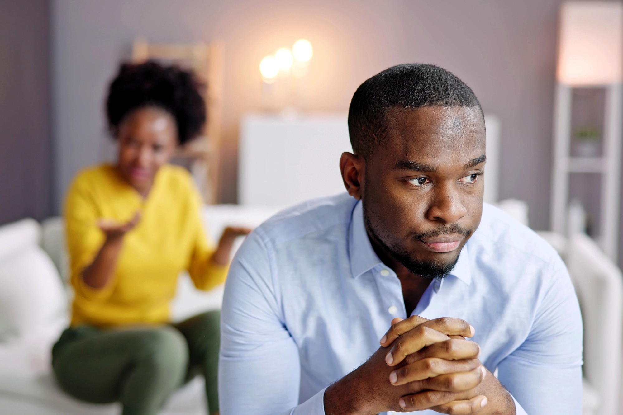 A man sits in the foreground looking thoughtful and concerned, with his hands clasped, while a woman in the background gestures emotionally, suggesting a serious conversation or disagreement between them.