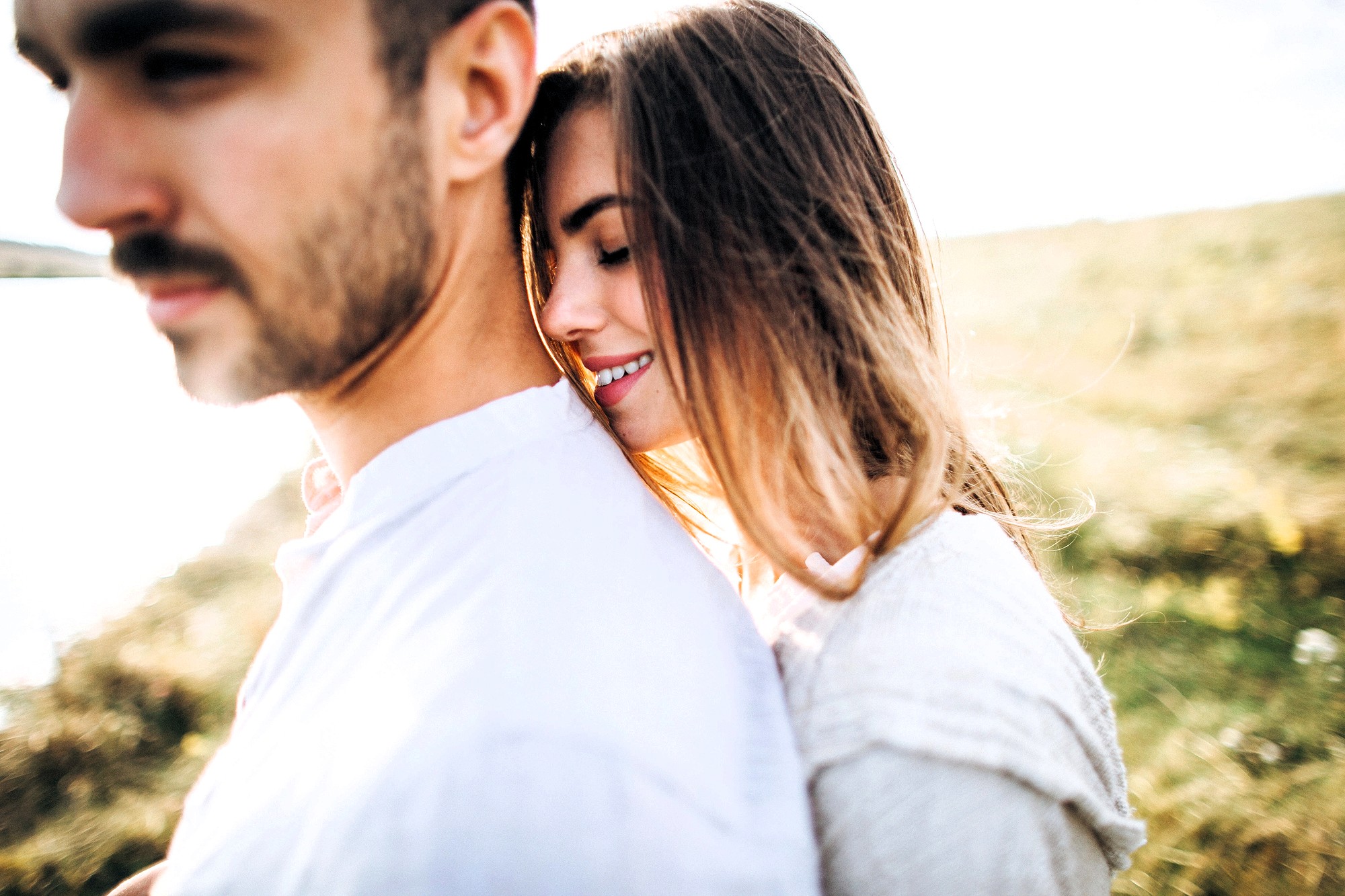 A woman with long hair leans her head on a man's shoulder, smiling with her eyes closed. The man, with short hair and a mustache, faces away from her. They stand outdoors in a sunlit, grassy field.