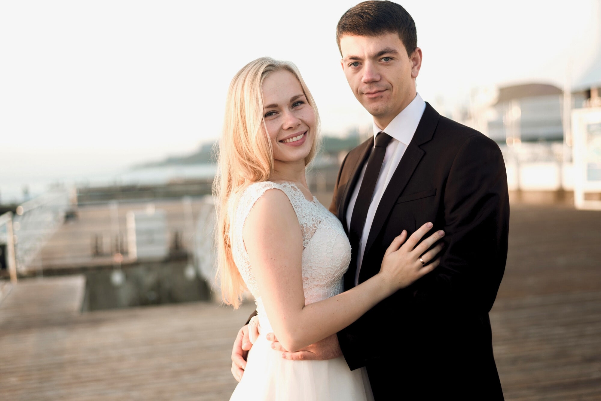 A smiling couple embraces on a wooden boardwalk. The woman wears a white sleeveless dress, and the man is in a black suit and tie. The background is bright and outdoors, with blurred railings and structures.