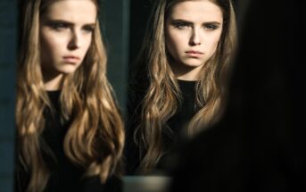 A young woman with long, wavy hair and a serious expression is looking at her reflection in a mirror. Soft light highlights her face, and both her and her reflection are visible in the image.