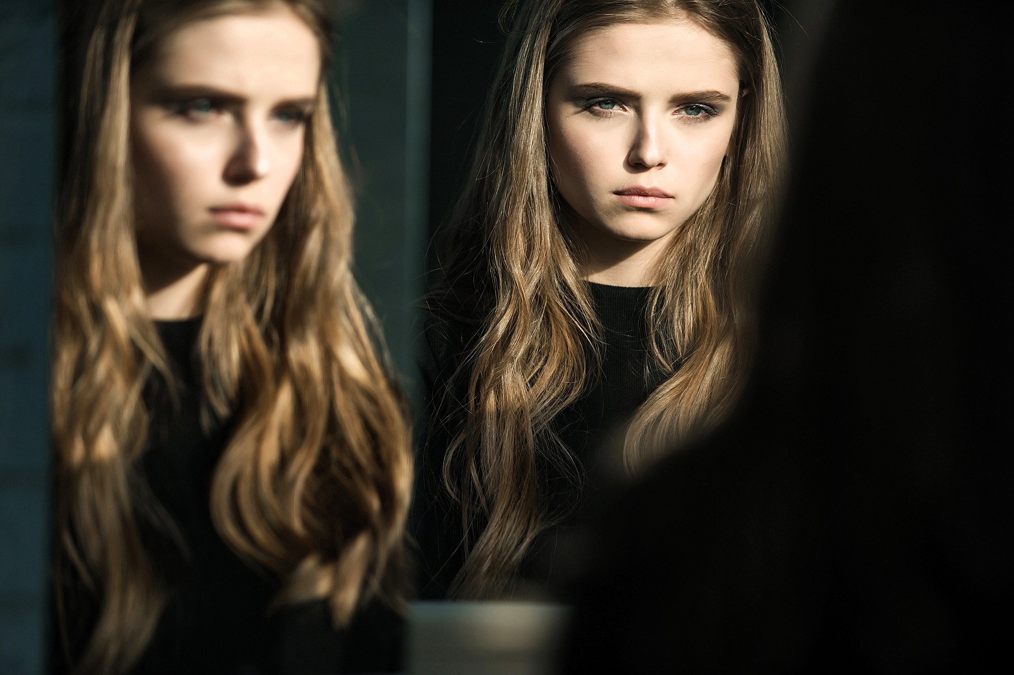 A young woman with long, wavy hair and a serious expression is looking at her reflection in a mirror. Soft light highlights her face, and both her and her reflection are visible in the image.