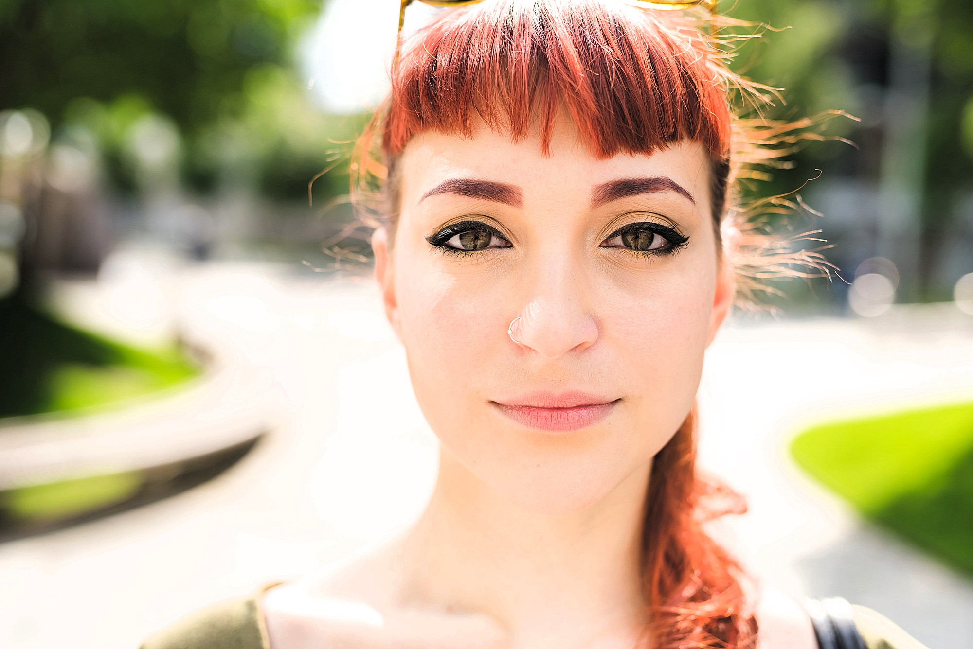 A young woman with red hair, winged eyeliner, and a nose ring looks directly at the camera outdoors on a sunny day, with a blurred background of green trees and a curved path.
