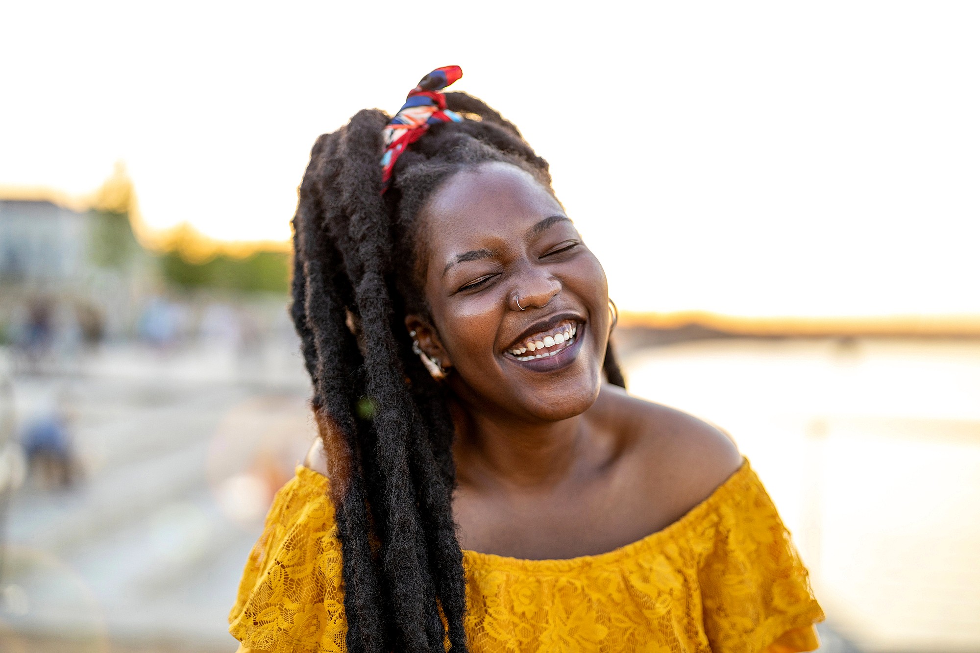 A woman with long, natural hair and a red headband smiles brightly while standing outdoors near water at sunset, wearing an off-the-shoulder yellow top.