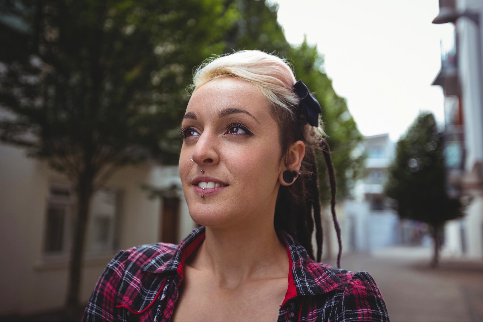 A person with light and dark hair styled in dreadlocks, wearing a plaid shirt and earrings, stands outdoors on a tree-lined street, looking slightly upward with a content expression.