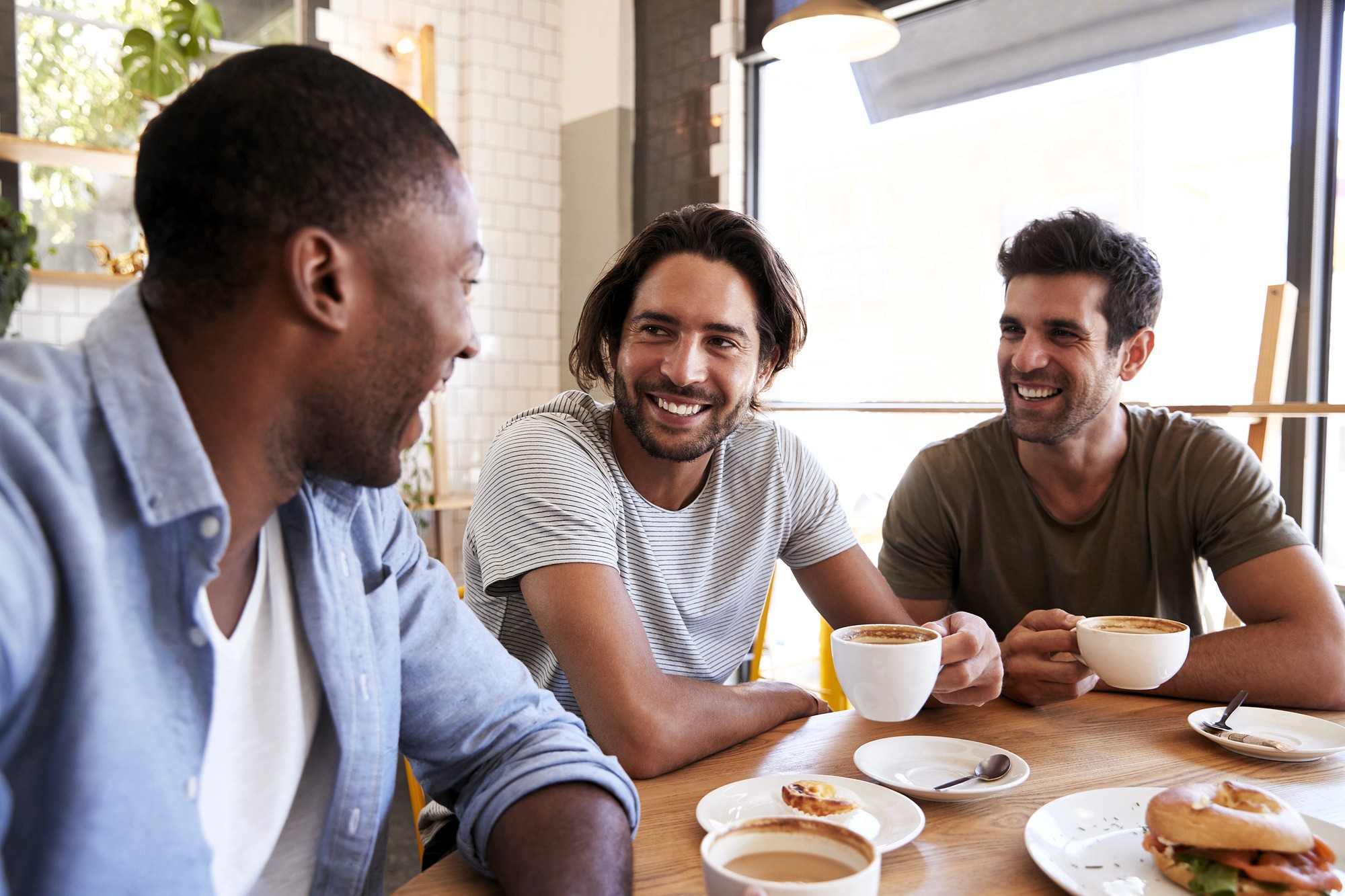 Three men sit at a café table, smiling and talking while holding coffee cups. Sunlight streams in through large windows, creating a warm and cheerful atmosphere.