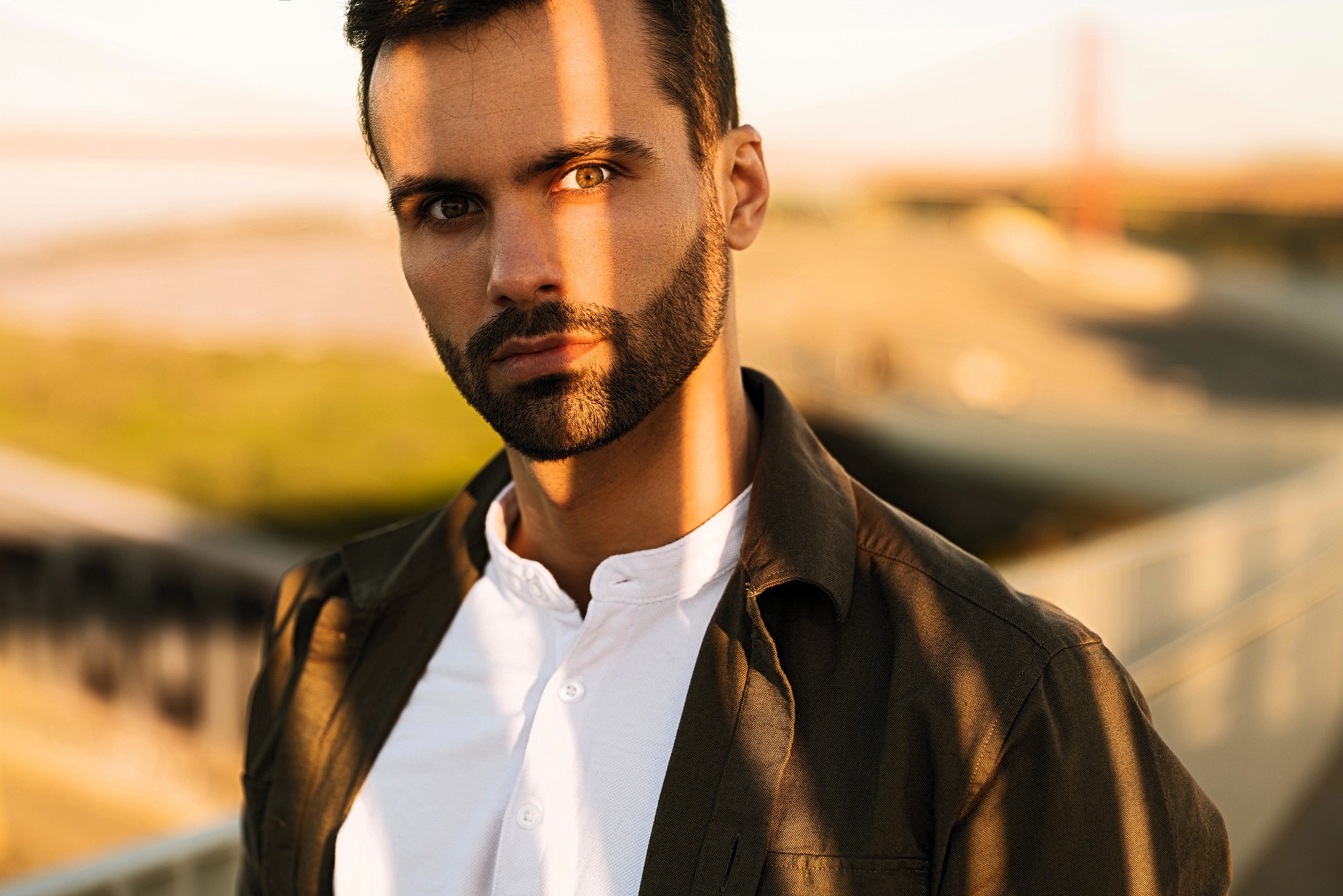 A man with dark hair and a beard stands outdoors, wearing a white shirt and a dark jacket. Sunlight casts a shadow across his face. The background is blurred, showing an outdoor walkway and distant structures.