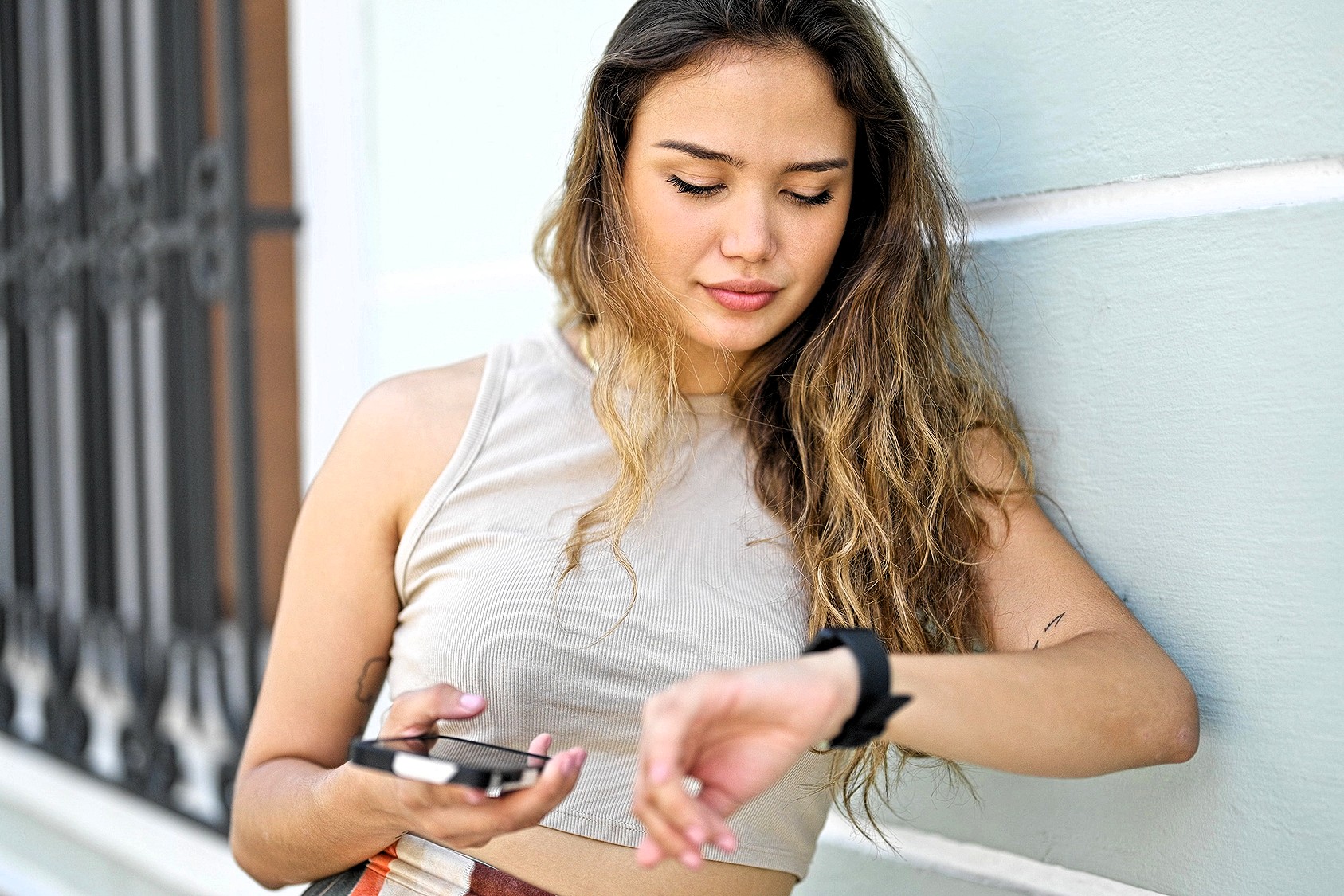 A young woman with long hair leans against a light-colored wall, holding a smartphone in one hand and looking down at her smartwatch on her wrist. She appears relaxed and focused.
