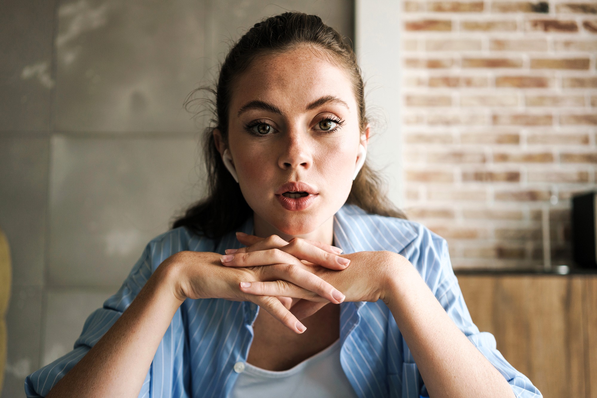 A young woman with light eyes and long brown hair, wearing a blue striped shirt, sits indoors with her hands clasped under her chin, looking attentively forward. A brick wall and tiled wall are in the background.