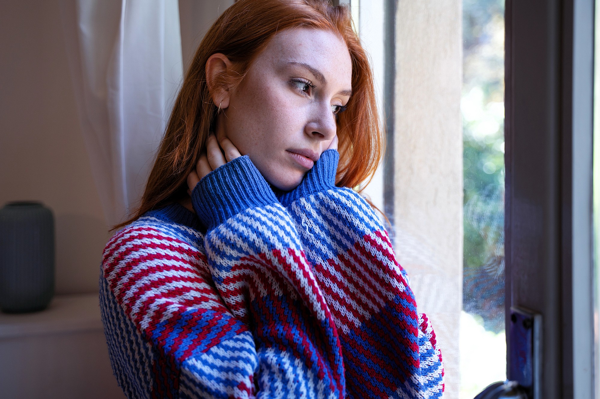 A young woman with long red hair and a colorful, striped sweater stands by a window, gazing outside with a thoughtful, pensive expression, her hands gently touching her neck.