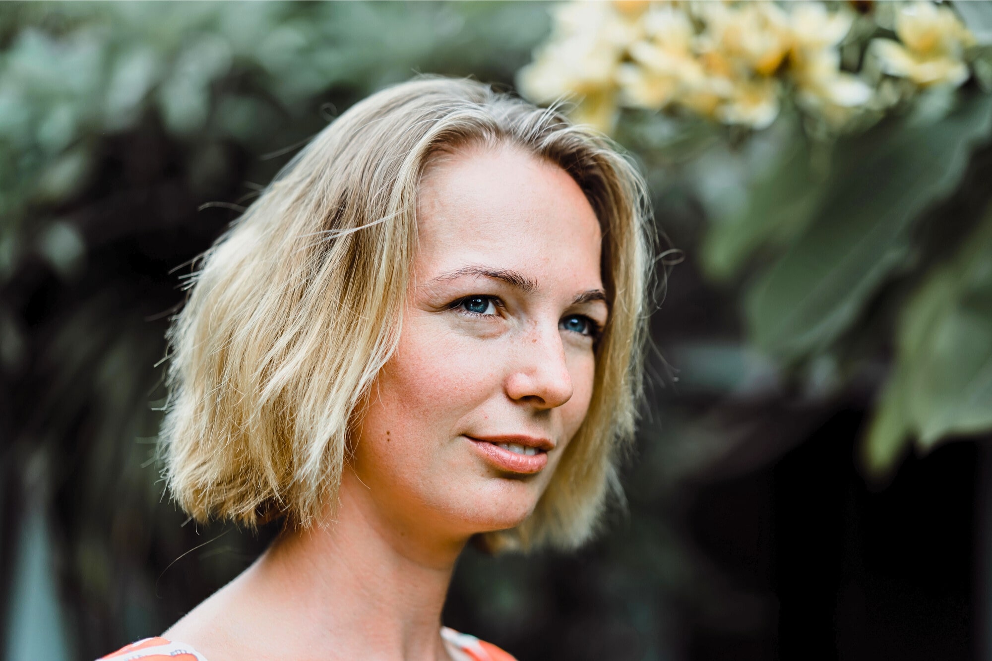 A woman with short blonde hair smiles softly outdoors, standing in front of green foliage and yellow flowers. The background is blurred, drawing attention to her face.