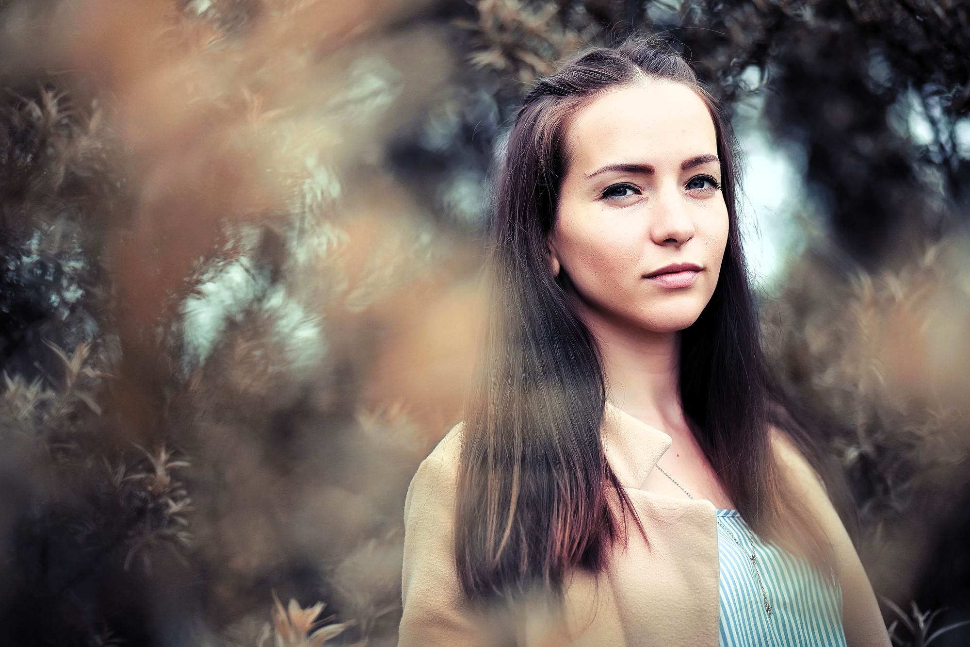 A young woman with long brown hair stands outdoors in front of blurry foliage, looking calmly at the camera. She wears a light-colored coat over a striped shirt. The image has a soft, natural, and slightly muted tone.