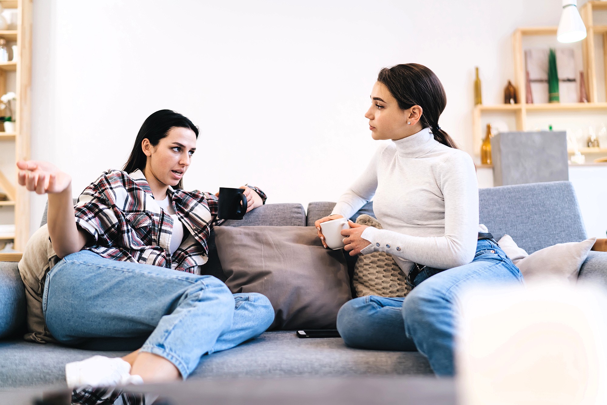 Two women sit on a couch having a serious conversation, each holding a mug. One gestures with her hand while speaking, and the other listens attentively. Both are dressed casually in jeans and tops.