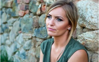 A woman with blonde hair and a green top sits against a stone wall outdoors, looking thoughtfully into the distance with a neutral expression.