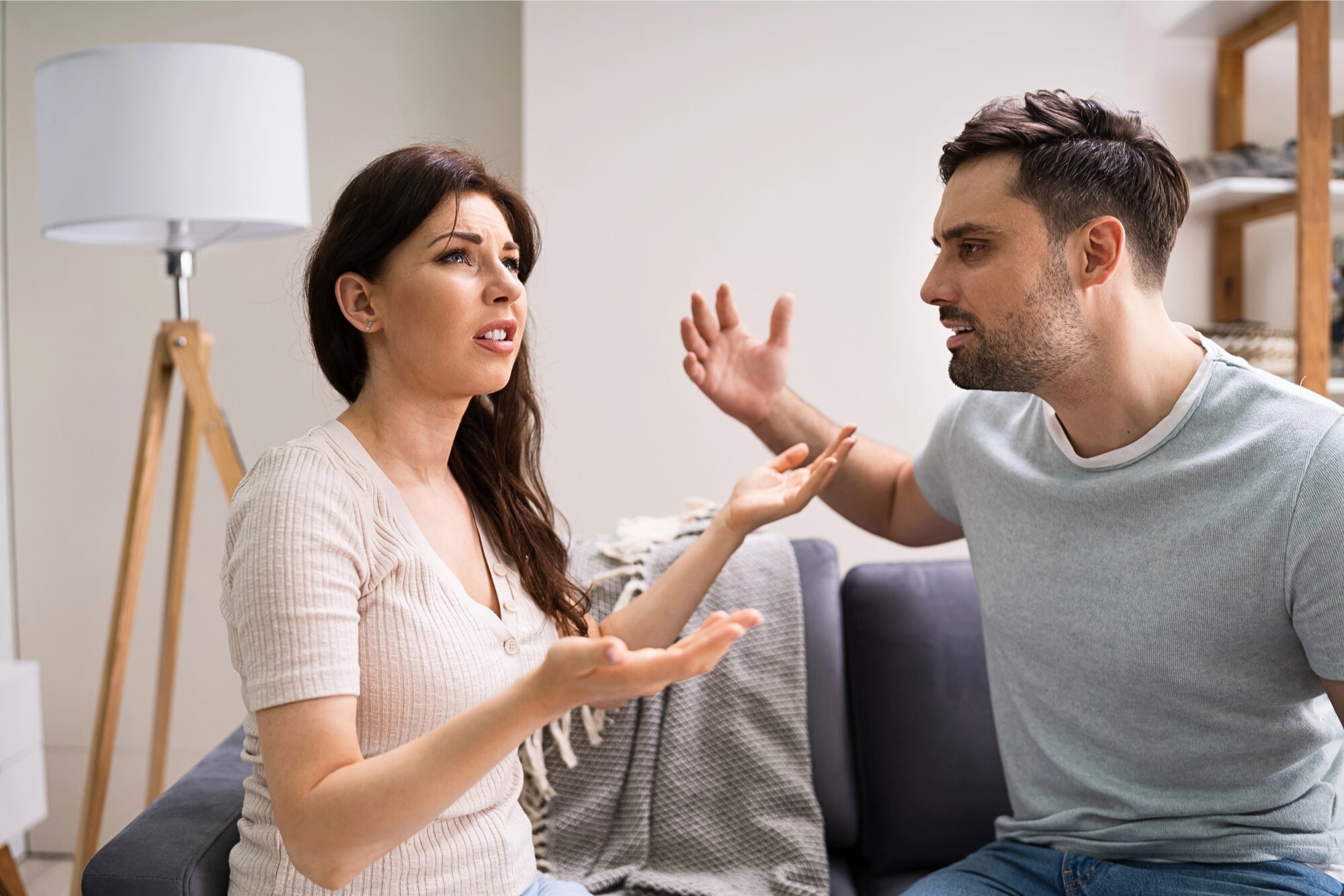 A woman and a man sit on a couch indoors, facing each other and appearing to have an intense argument. Both have raised hands and frustrated facial expressions. A lamp and shelving are visible in the background.
