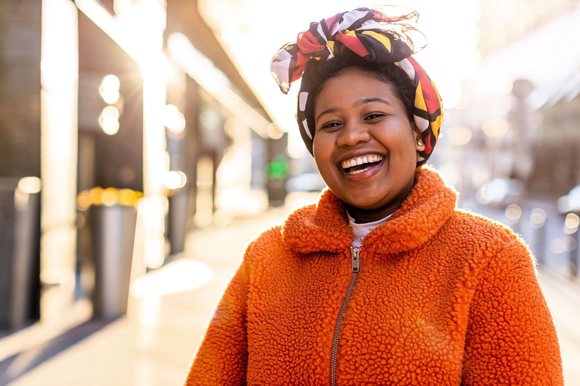 A woman wearing an orange fleece jacket and a colorful headscarf smiles brightly while standing outdoors in a sunlit urban setting.