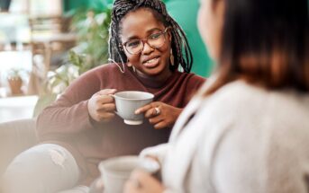 Two women sit indoors, holding mugs and having a conversation. One woman, wearing glasses and a brown sweater, smiles while speaking to the other, who is slightly out of focus in the foreground.