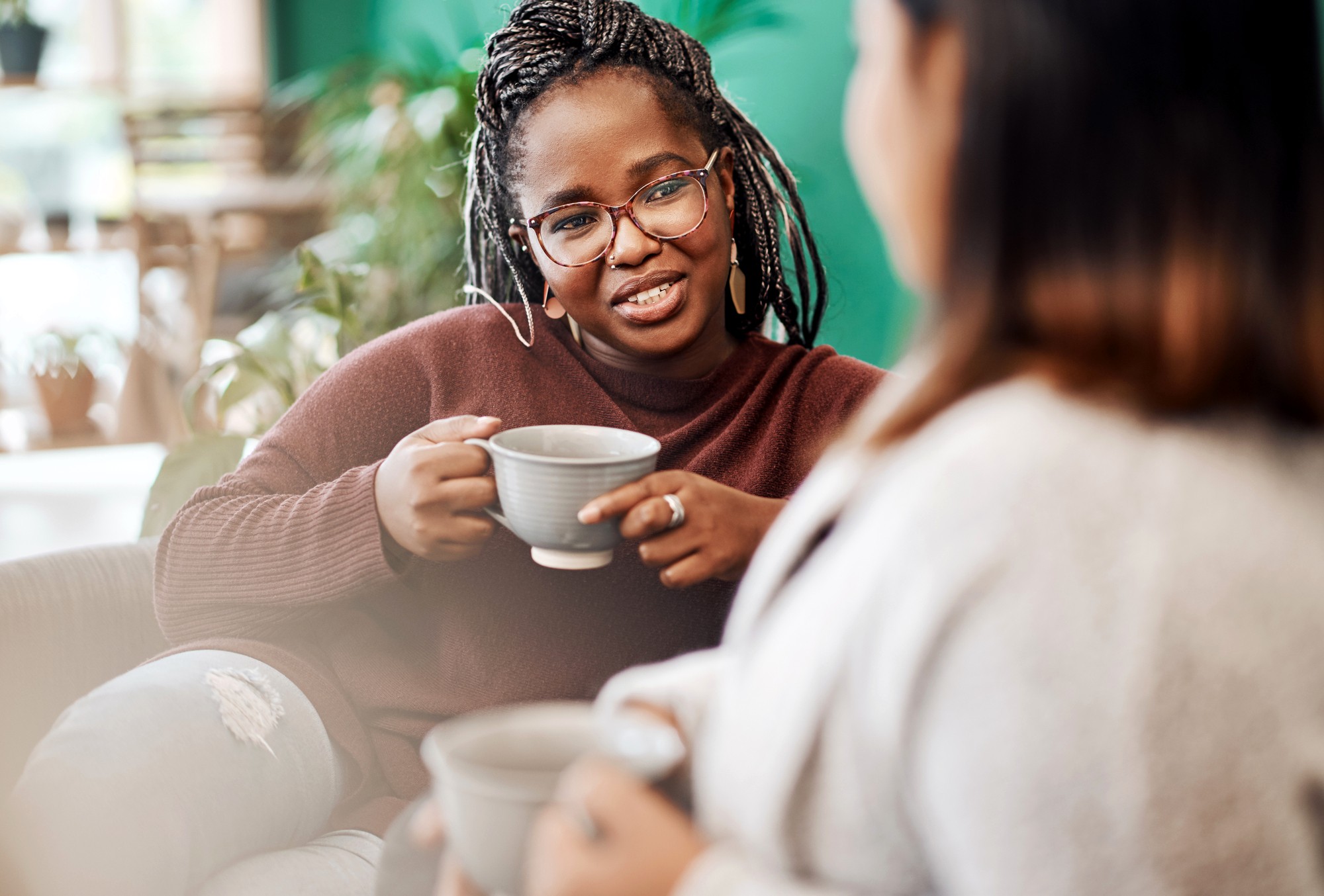 Two women sit indoors, holding mugs and having a conversation. One woman, wearing glasses and a brown sweater, smiles while speaking to the other, who is slightly out of focus in the foreground.