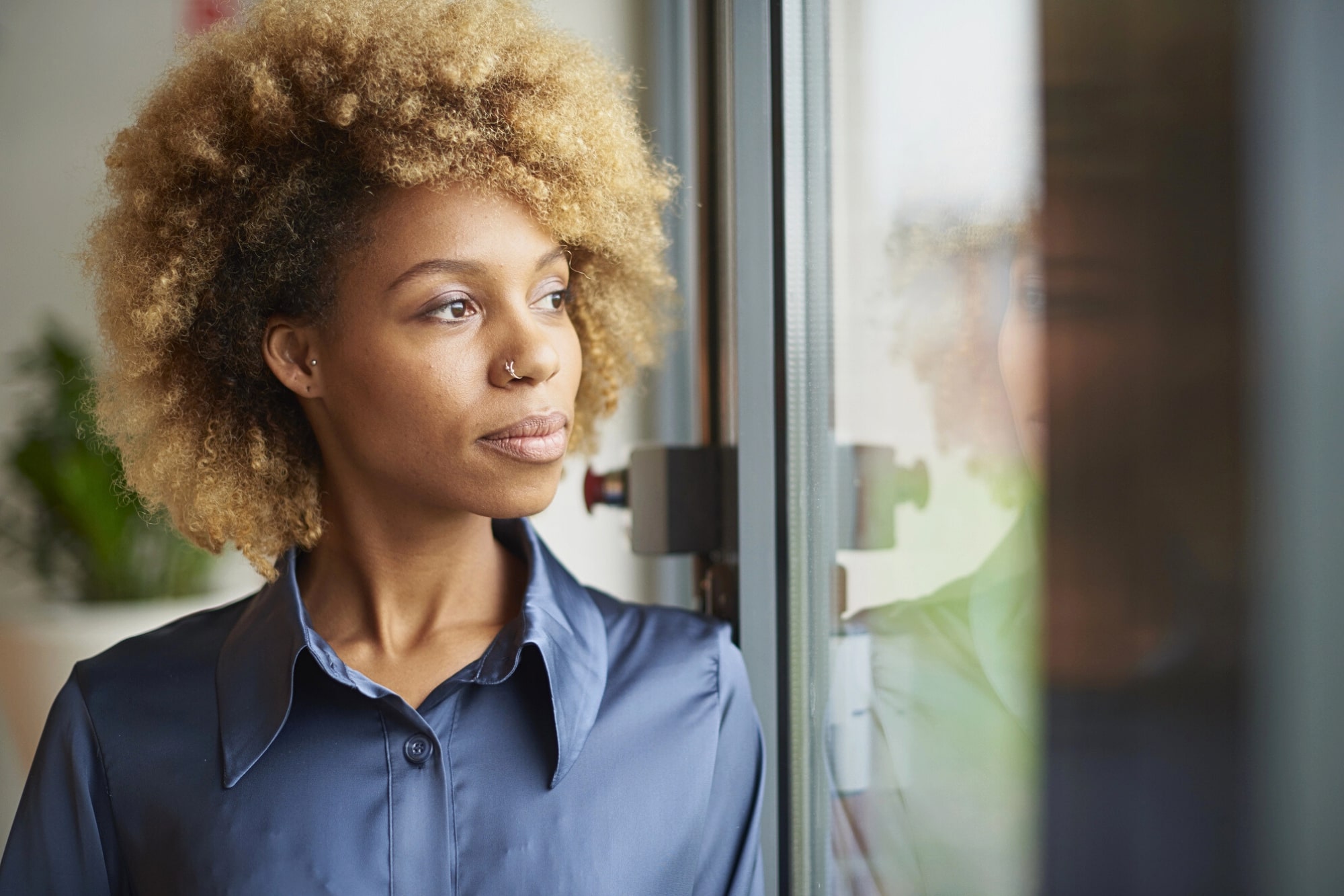 A woman with curly blonde hair and a nose ring gazes thoughtfully out of a window. She is wearing a shiny blue button-up shirt, and soft natural light illuminates her face. A blurred plant is visible in the background.