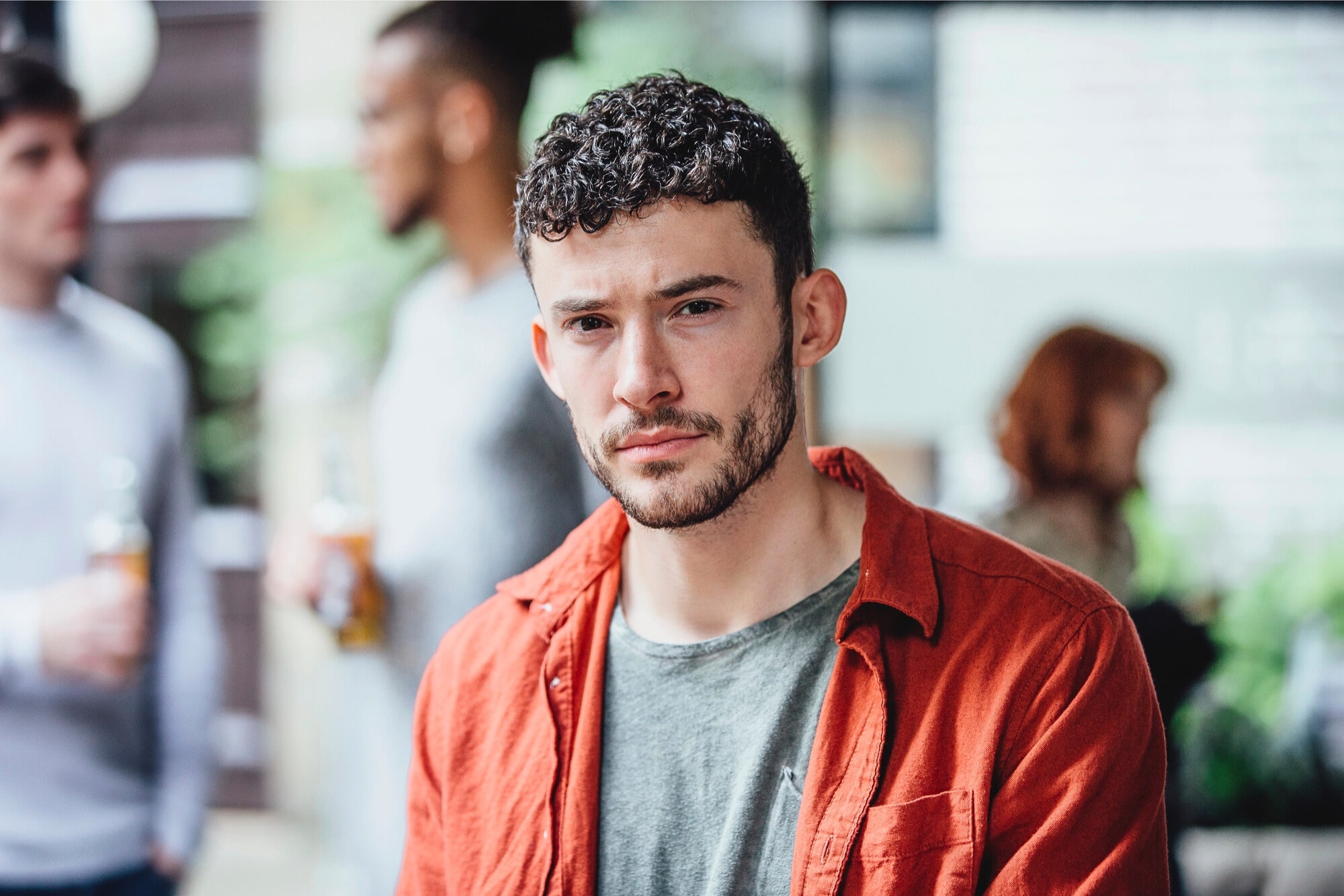 A man with short curly hair and a trimmed beard, wearing a red shirt over a gray t-shirt, looks at the camera. Several people are blurred in the background, standing and talking outdoors.