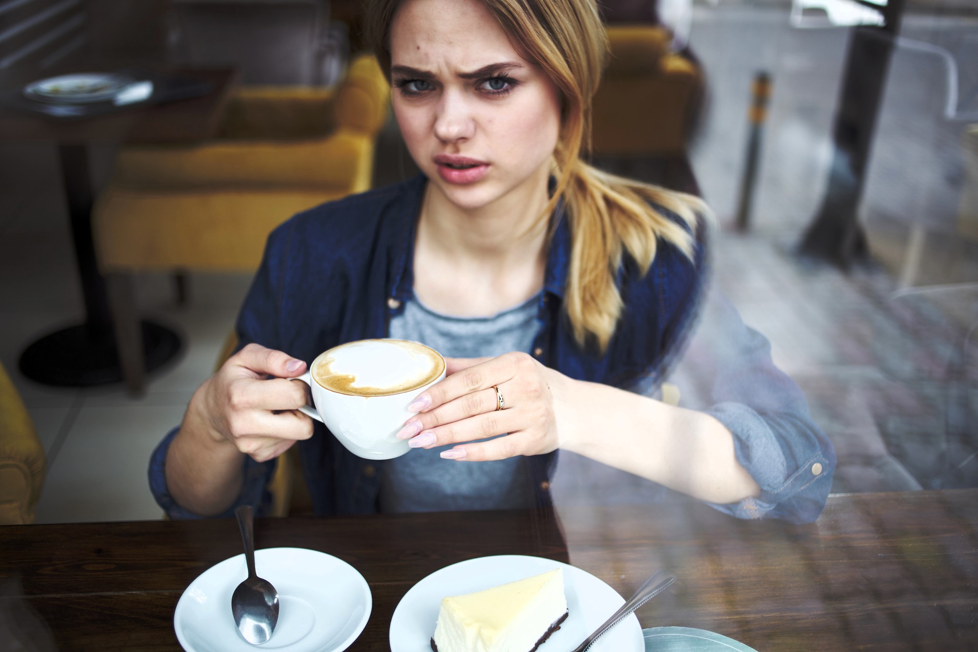 A woman sits at a café table, holding a cup of coffee and looking confused or displeased. A plate with a slice of cake, a fork, and a spoon are on the table in front of her. The photo is taken through a window.