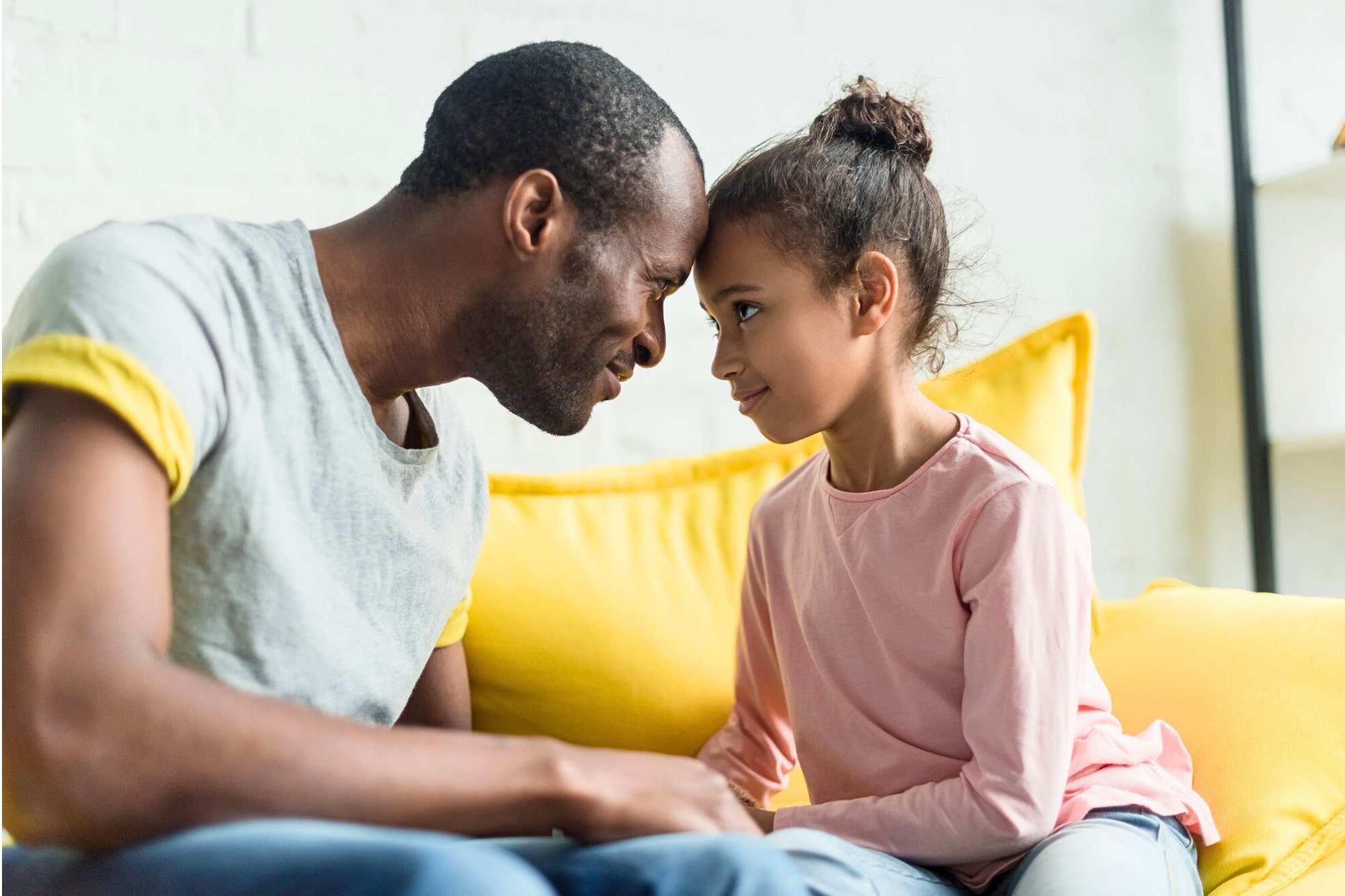 A man and a young girl sit on a yellow couch, touching foreheads and smiling warmly at each other, showing a close and affectionate bond.