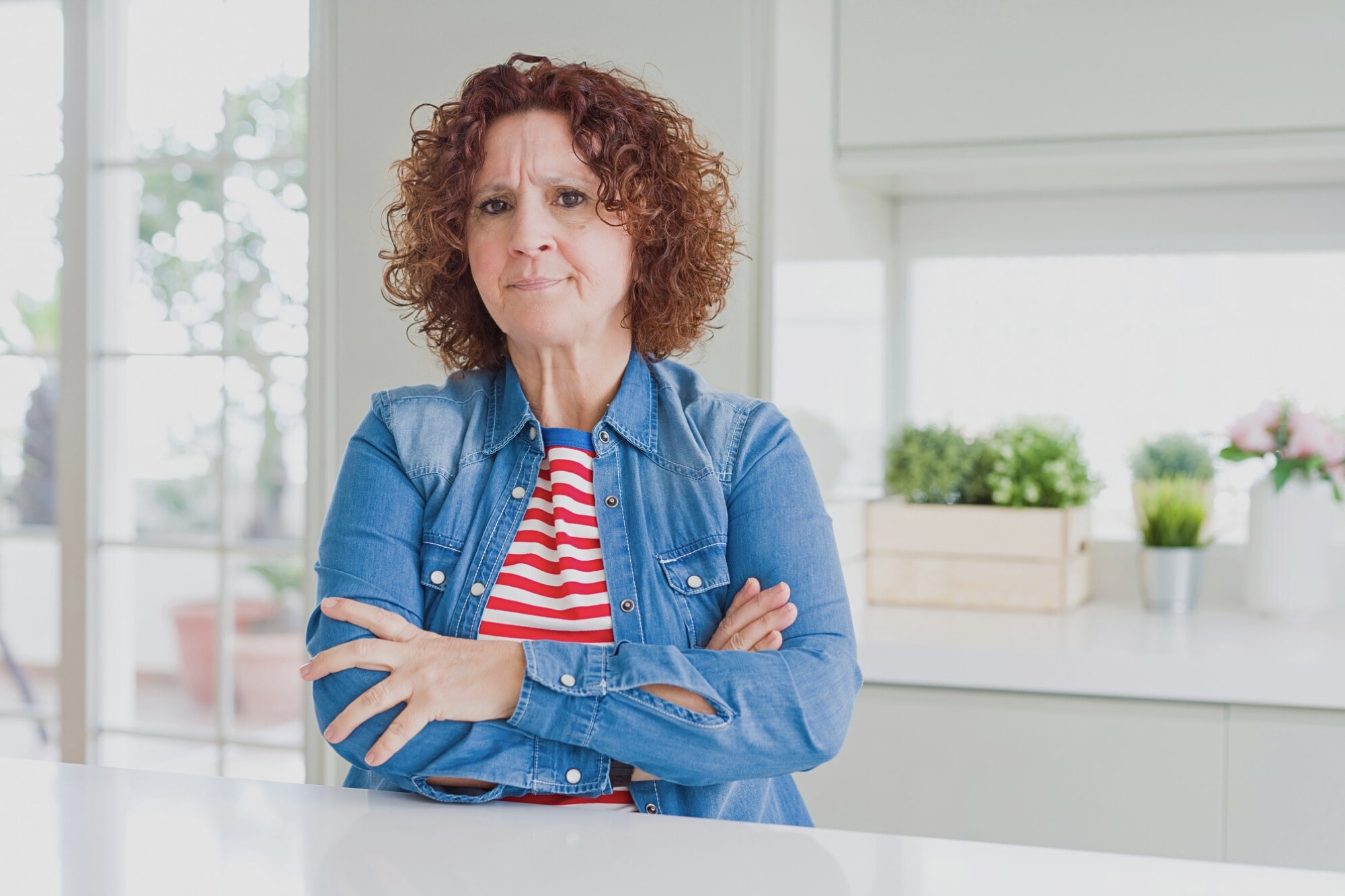 A woman with curly brown hair, wearing a denim jacket over a red and white striped shirt, stands in a bright kitchen with her arms crossed, looking serious. Green plants are visible on the counter in the background.