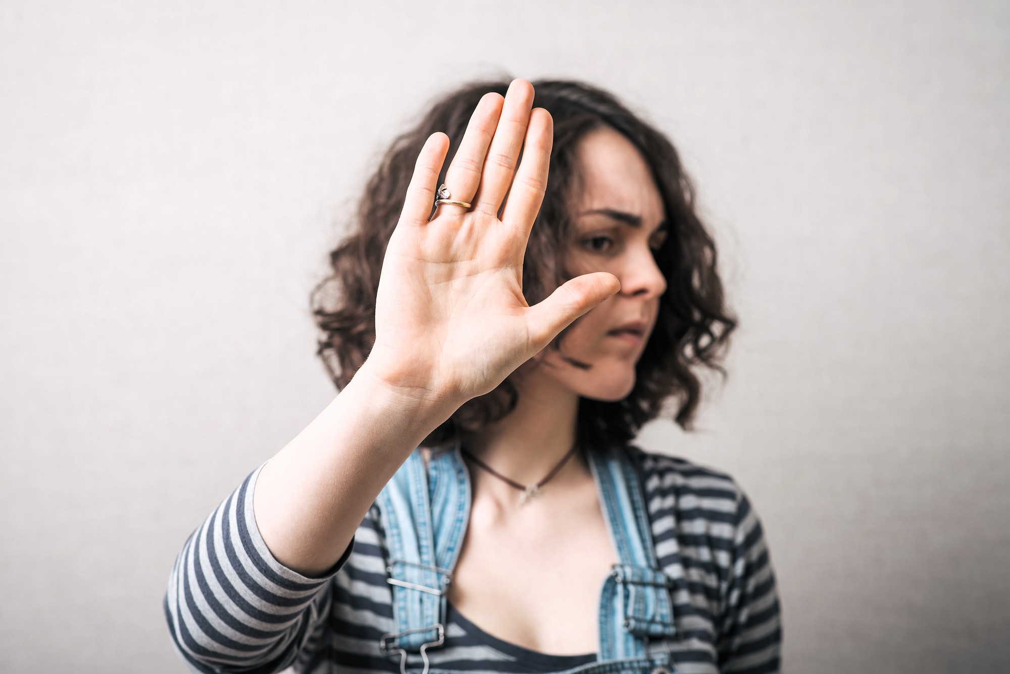 A woman with curly hair wearing a striped shirt and denim overalls holds up her hand in front of her face as if signaling "stop" or "no," with a serious expression and looking to the side.