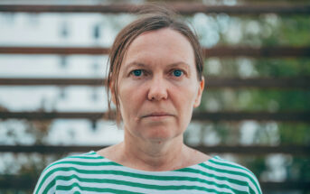 A woman with light skin and straight brown hair pulled back is wearing a green and white striped shirt. She stands outdoors in front of a blurred background of horizontal wooden slats and greenery, looking directly at the camera.