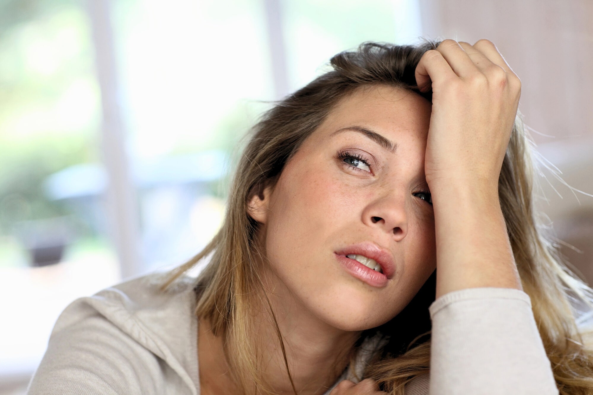 A young woman with long blonde hair rests her head on her hand, looking tired or stressed. She is indoors with a blurred background, wearing a light-colored top.