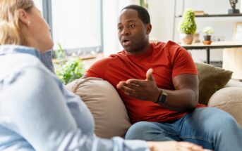 A man in a red shirt sits on a couch, gesturing with his hand as he talks to a woman in a light blue shirt. They appear to be having a serious conversation in a bright living room.