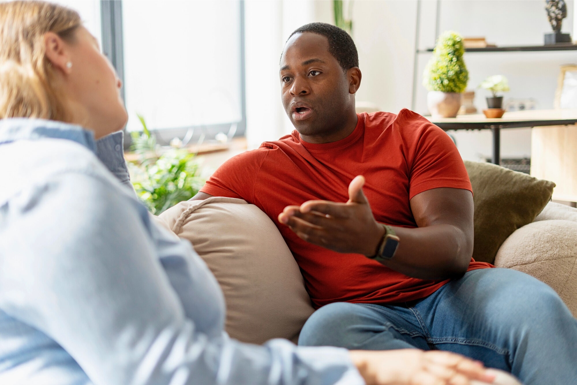 A man in a red shirt sits on a couch, gesturing with his hand as he talks to a woman in a light blue shirt. They appear to be having a serious conversation in a bright living room.