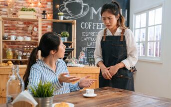 A woman sitting at a table in a café gestures with concern toward a waitress in an apron, who stands looking worried. A chalkboard menu and shelves with jars are visible in the background.