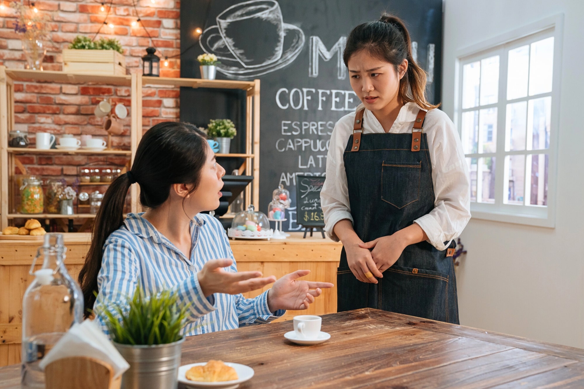 A woman sitting at a table in a café gestures with concern toward a waitress in an apron, who stands looking worried. A chalkboard menu and shelves with jars are visible in the background.