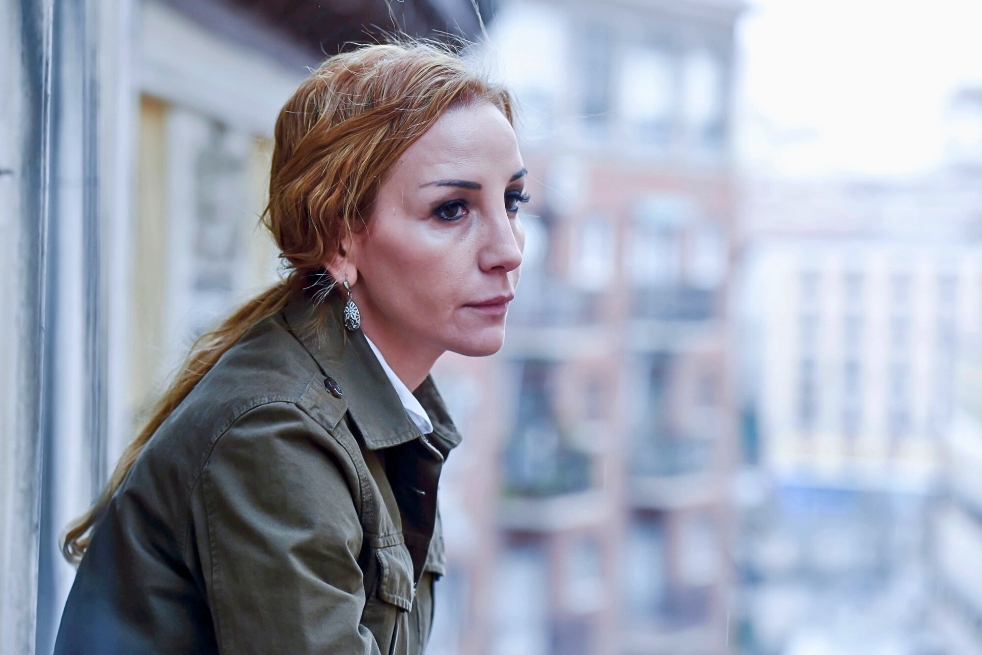 A woman with reddish-brown hair in a ponytail and wearing a green jacket leans on a balcony railing, looking thoughtfully into the distance, with blurred buildings in the background.