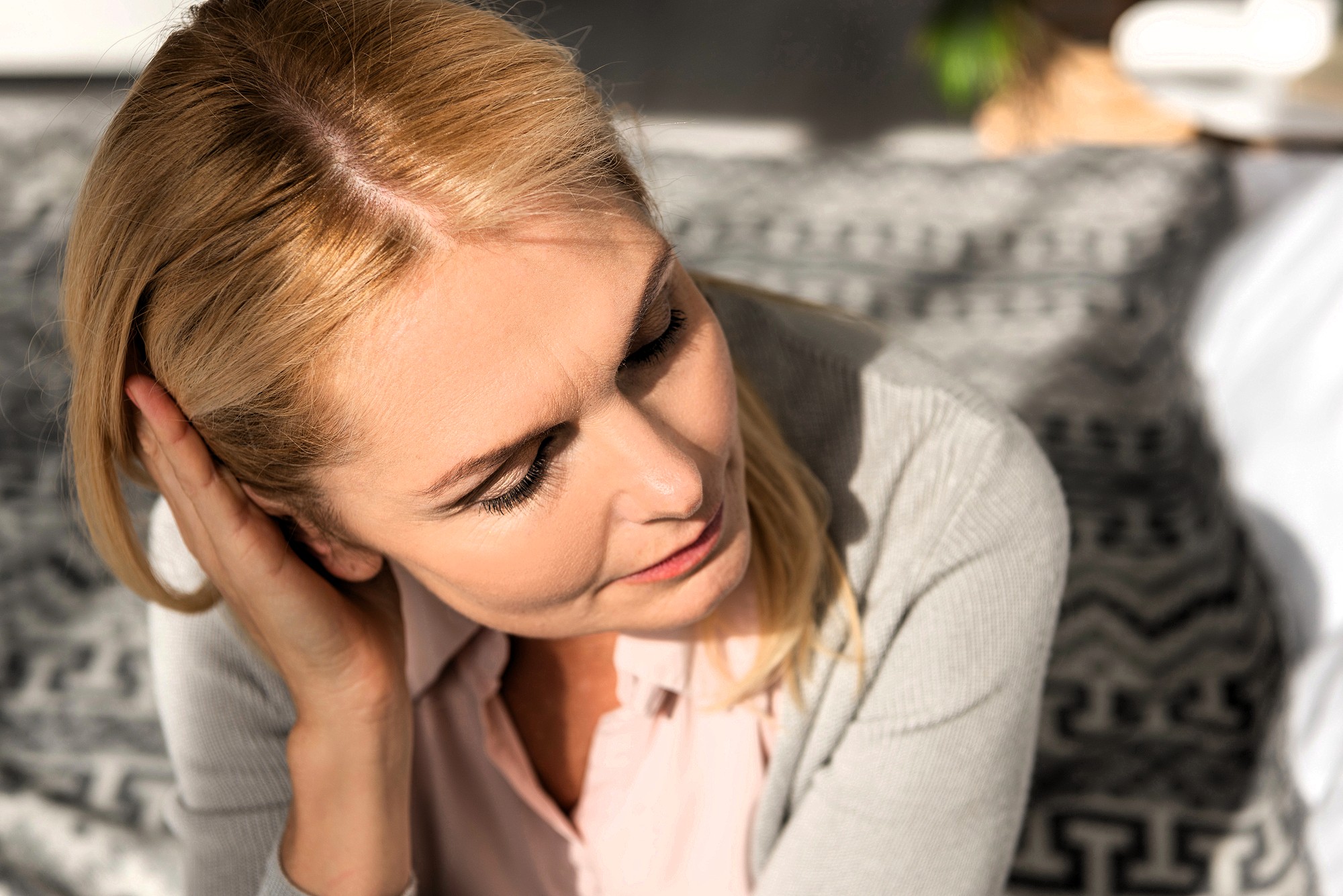 A middle-aged woman with blonde hair looks down thoughtfully while touching her hair, sitting indoors on a patterned blanket in soft natural light.