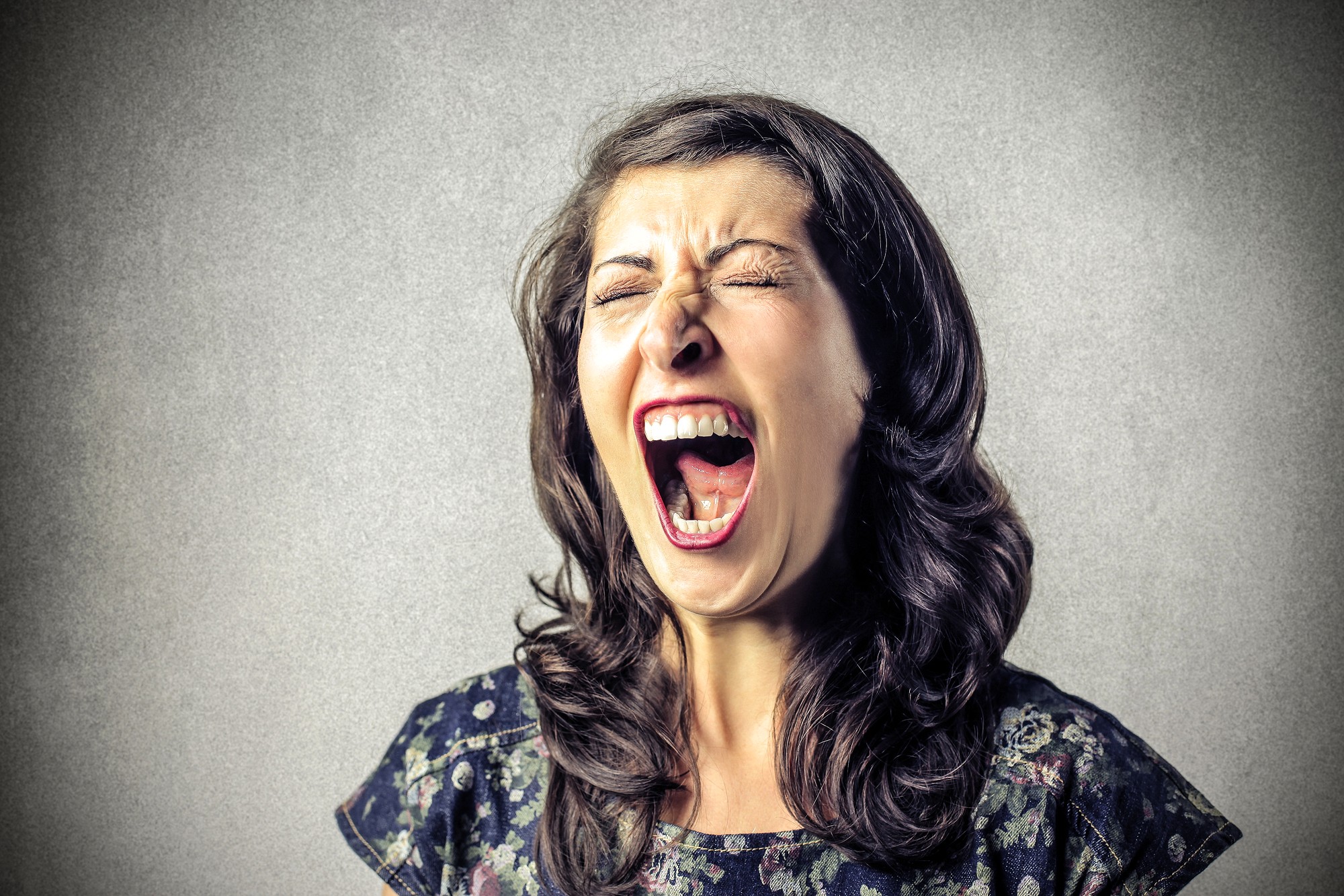 A woman with long dark hair, wearing a patterned top, is standing against a gray background. Her eyes are closed and her mouth is wide open, as if she is screaming or shouting.