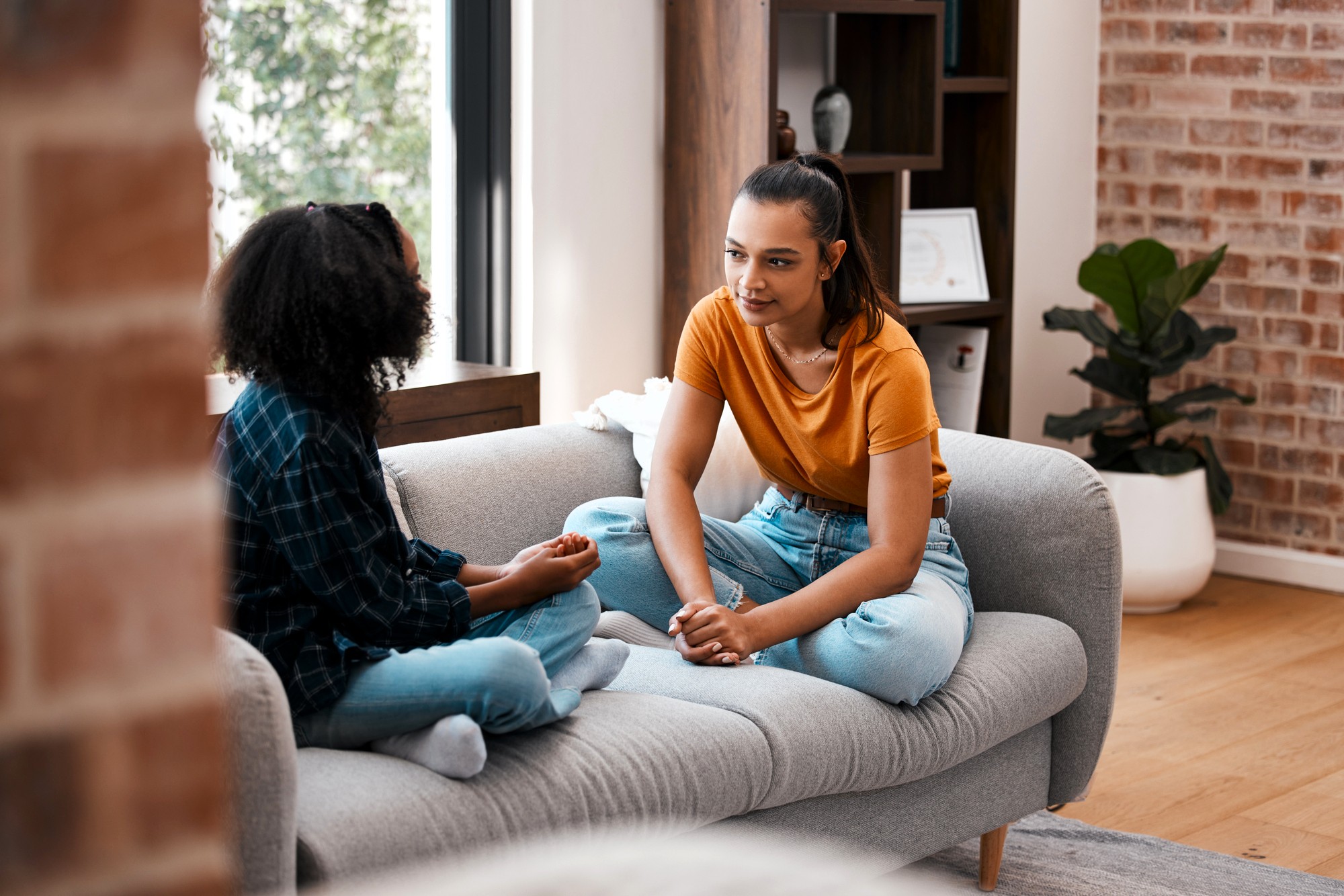 Two young women sit on a gray couch in a cozy living room, facing each other and having a serious conversation. One wears an orange shirt and jeans, the other a plaid shirt and jeans. Natural light fills the space.