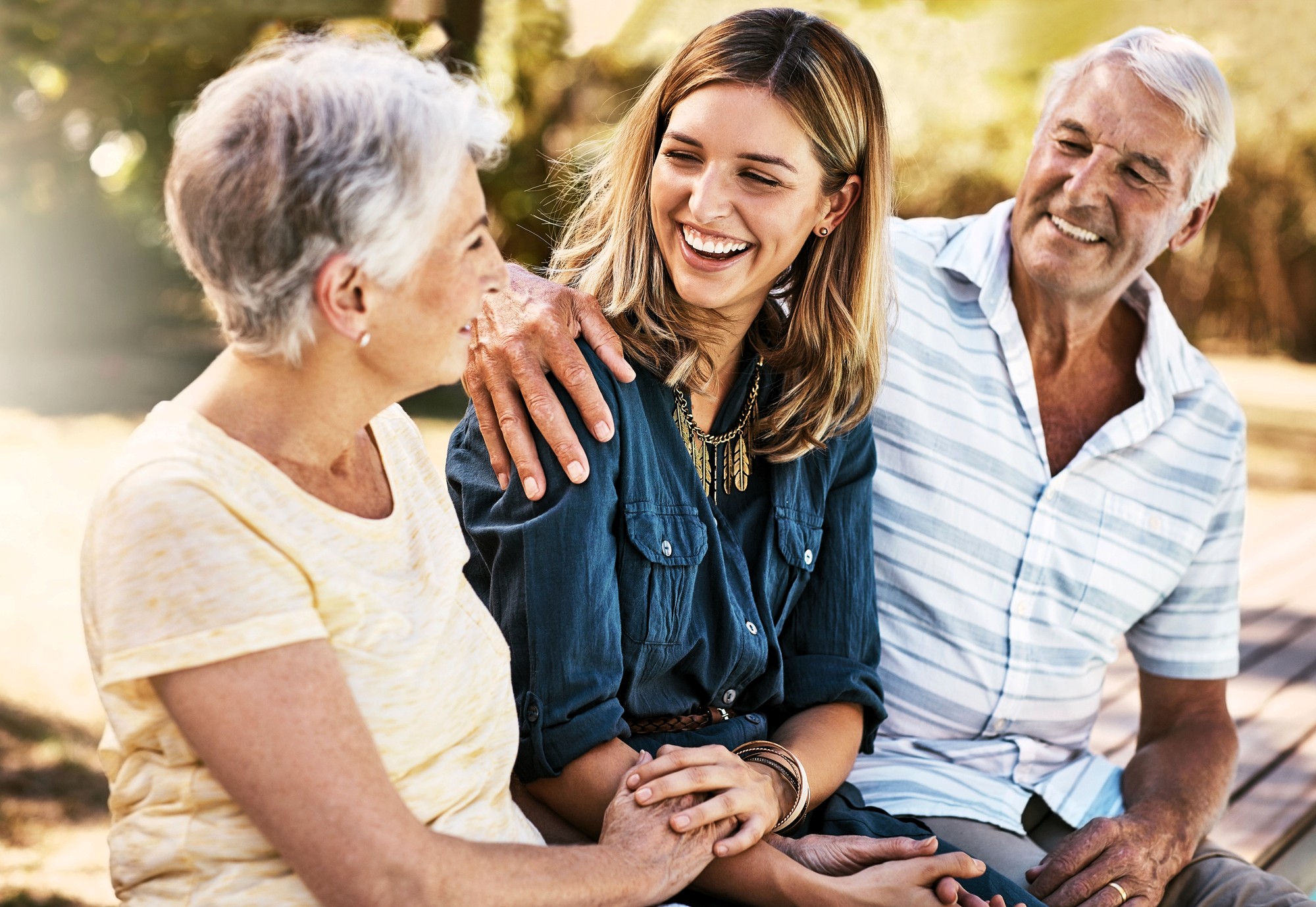 A young woman sits outdoors on a bench, smiling and holding hands with an older woman, while an older man with his arm around the woman looks on, all appearing happy and relaxed together.