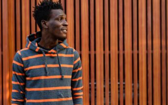 A person wearing a gray and orange striped hoodie stands in front of a wooden slatted wall, looking to the side and smiling slightly.