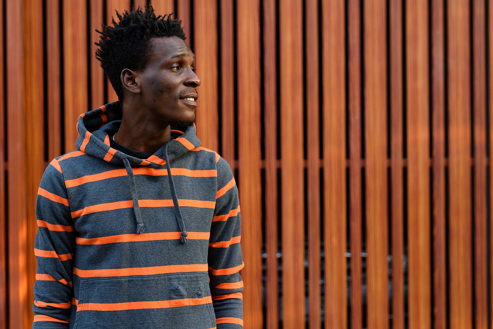 A person wearing a gray and orange striped hoodie stands in front of a wooden slatted wall, looking to the side and smiling slightly.