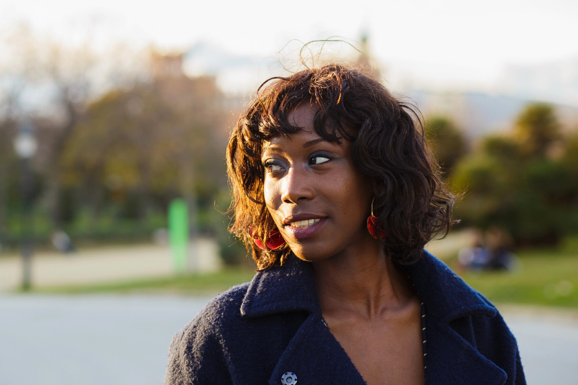 A woman with short, curly hair wearing a dark coat and red earrings stands outdoors, looking to the side and smiling, with a blurred park and trees in the background.