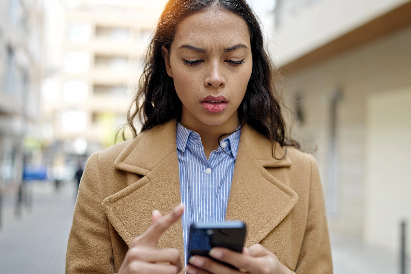 A woman with long dark hair wearing a tan coat and striped shirt looks at her smartphone with a concerned expression while standing outdoors on a city street.