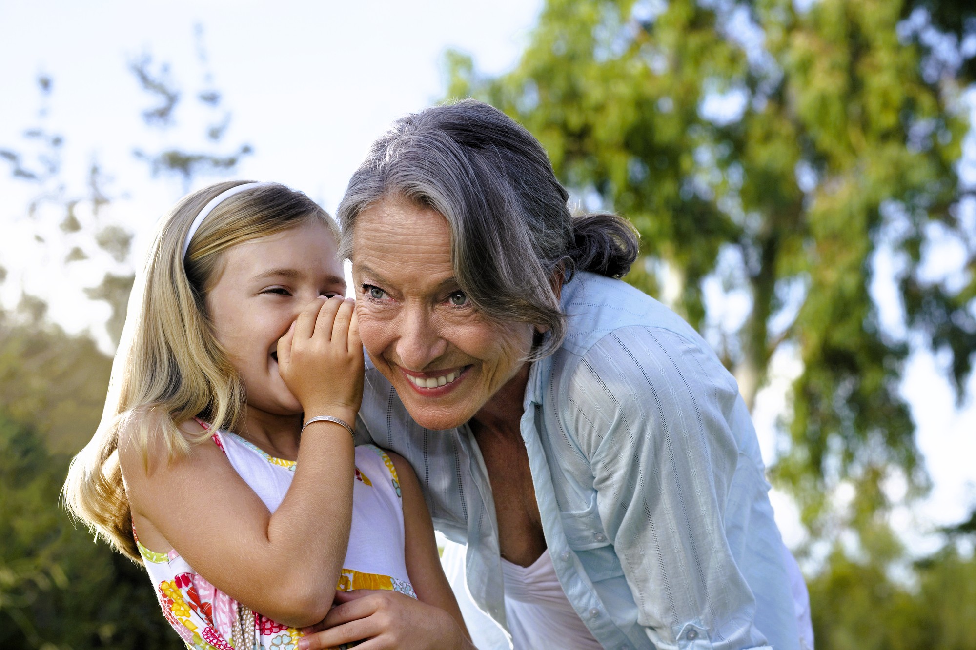 A young girl whispers into an older woman's ear, both smiling and enjoying a moment together outdoors, with green trees and sunlight in the background.