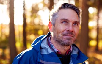 A middle-aged man with gray hair and a beard smiles softly while standing outdoors in a sunlit forest. He is wearing a blue jacket over a plaid shirt, with sunlight filtering through the trees behind him.