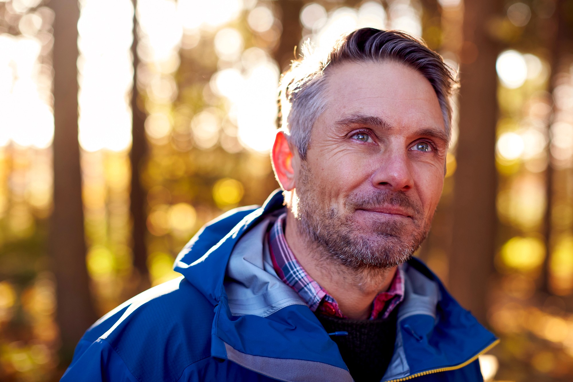A middle-aged man with gray hair and a beard smiles softly while standing outdoors in a sunlit forest. He is wearing a blue jacket over a plaid shirt, with sunlight filtering through the trees behind him.