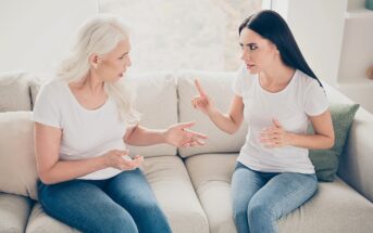 Two women sitting on a beige couch are having an intense conversation; the older woman gestures with open hands while the younger woman raises her index finger as if making a point. Both wear white tops and blue jeans.