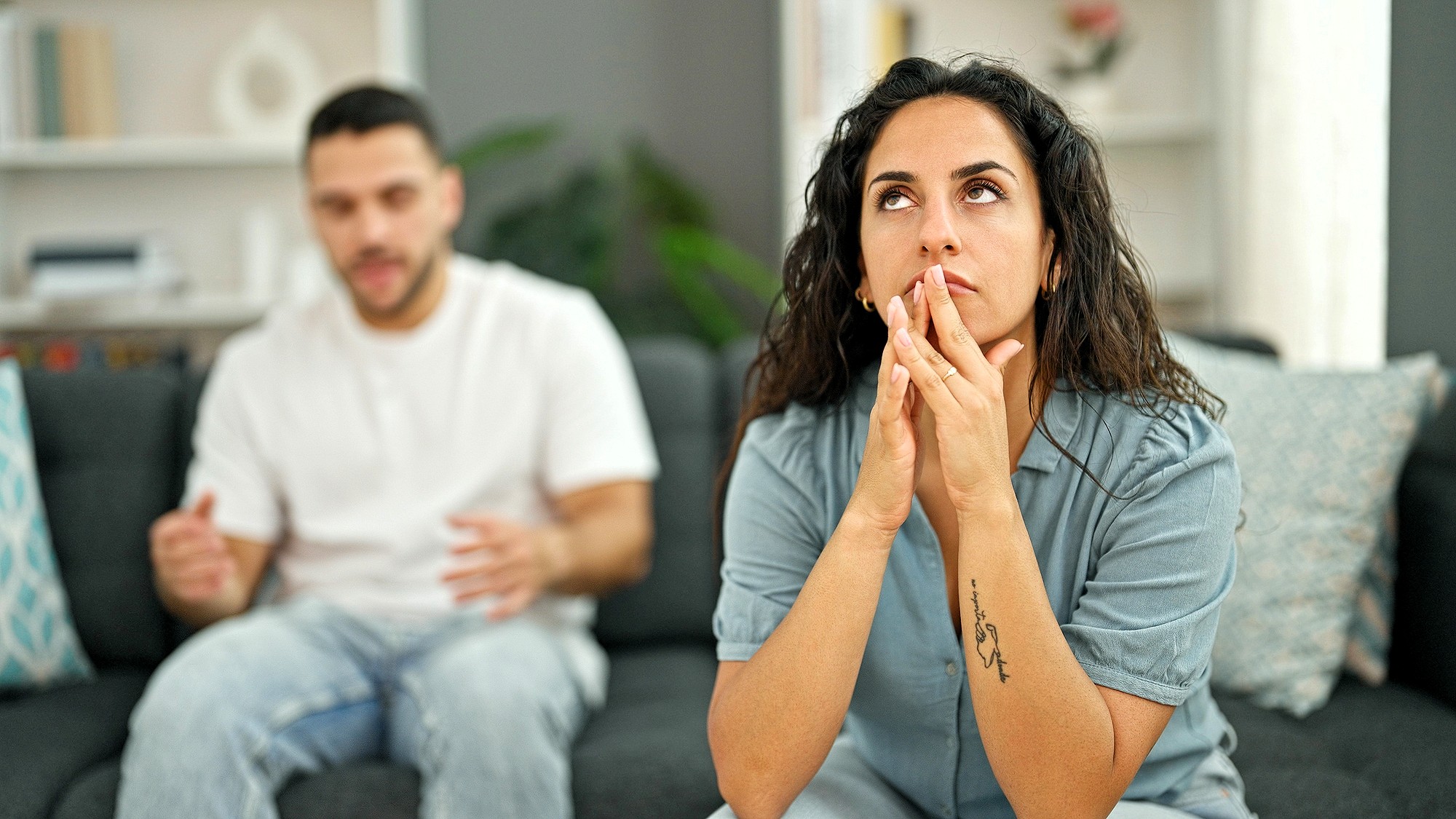 A woman sits on a couch looking frustrated and thoughtful, with her hands near her face, while a man in the background appears to be talking to her, looking concerned.