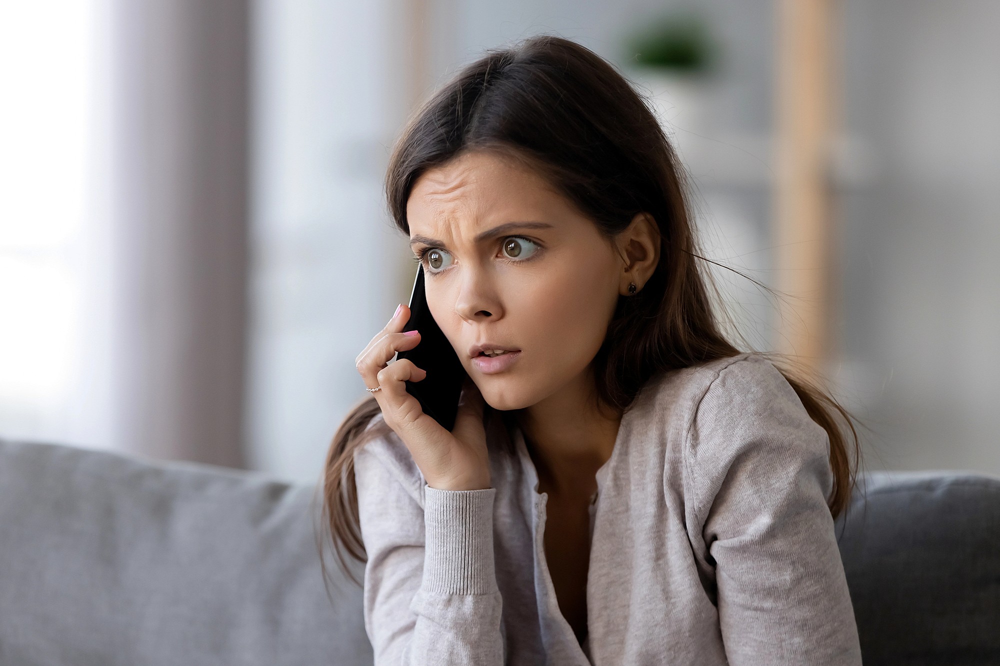 A young woman with long brown hair, wearing a light sweater, sits on a couch, looking worried while talking on a smartphone. Her brow is furrowed and her expression appears concerned or anxious.